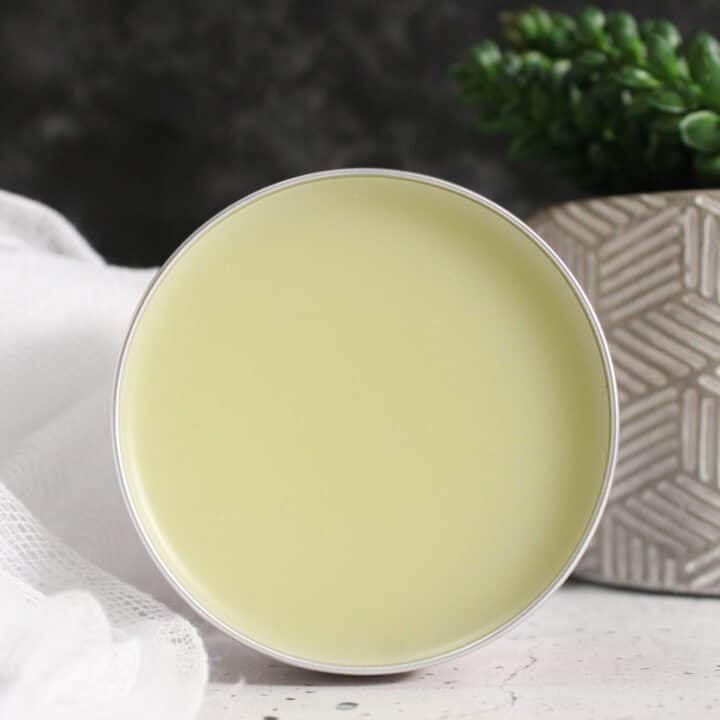 Pale yellow homemade bug bite balm in metal tin with white cloth and fresh herbs