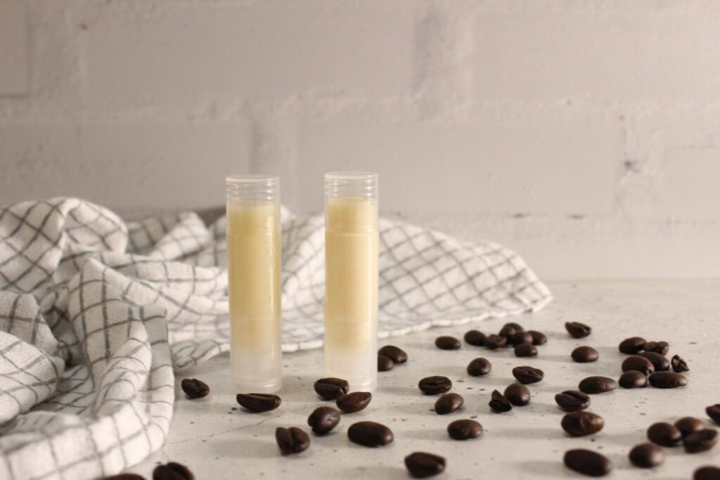 Two tubes of coffee lip balm standing upright, styled with coffee beans and a soft cloth in the background.