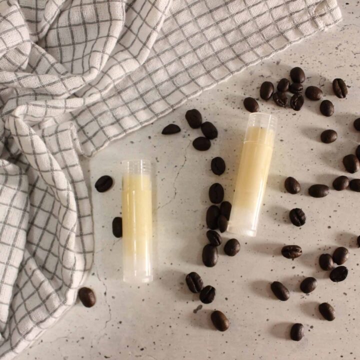 Two tubes of coffee lip balm arranged flat with coffee beans scattered around on a light surface, styled with a checkered cloth.