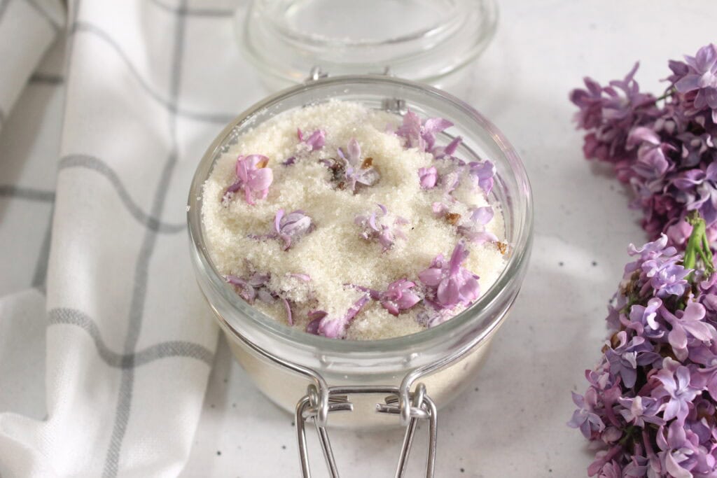 Lavender sugar in a glass jar with purple flowers nearby.