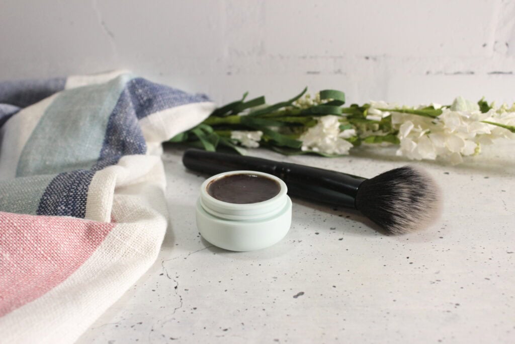 Open jar of homemade natural bronzer with a dark cream product, placed beside a makeup brush on a white surface, with fabric and white flowers in the background.