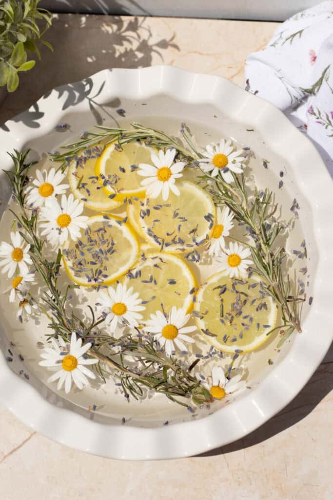 White bowl with water, lemon slices, daisies, lavender buds, and rosemary sprigs.