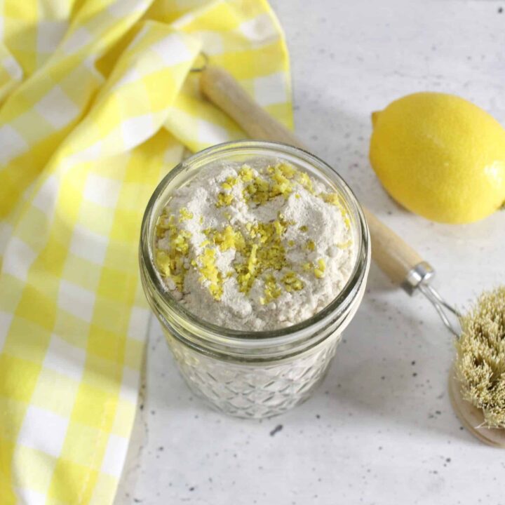 Glass jar filled with lemon sink cleaner powder topped with lemon zest, placed beside a natural scrub brush, a whole lemon, and a yellow checkered cloth on a light countertop.