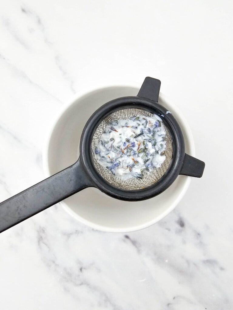 Strainer with honey lavender ice cream mixture over a white bowl on marble surface.