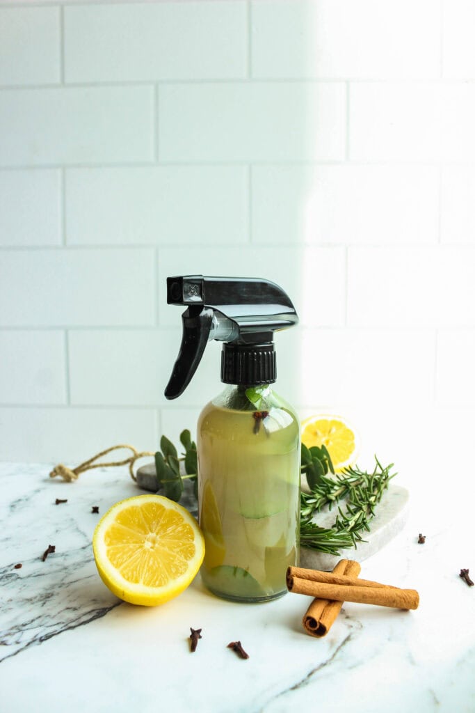 Homemade herbal cleaner in a glass spray bottle with cinnamon and rosemary inside, surrounded by lemon halves, whole cloves, and fresh herbs on a marble surface.