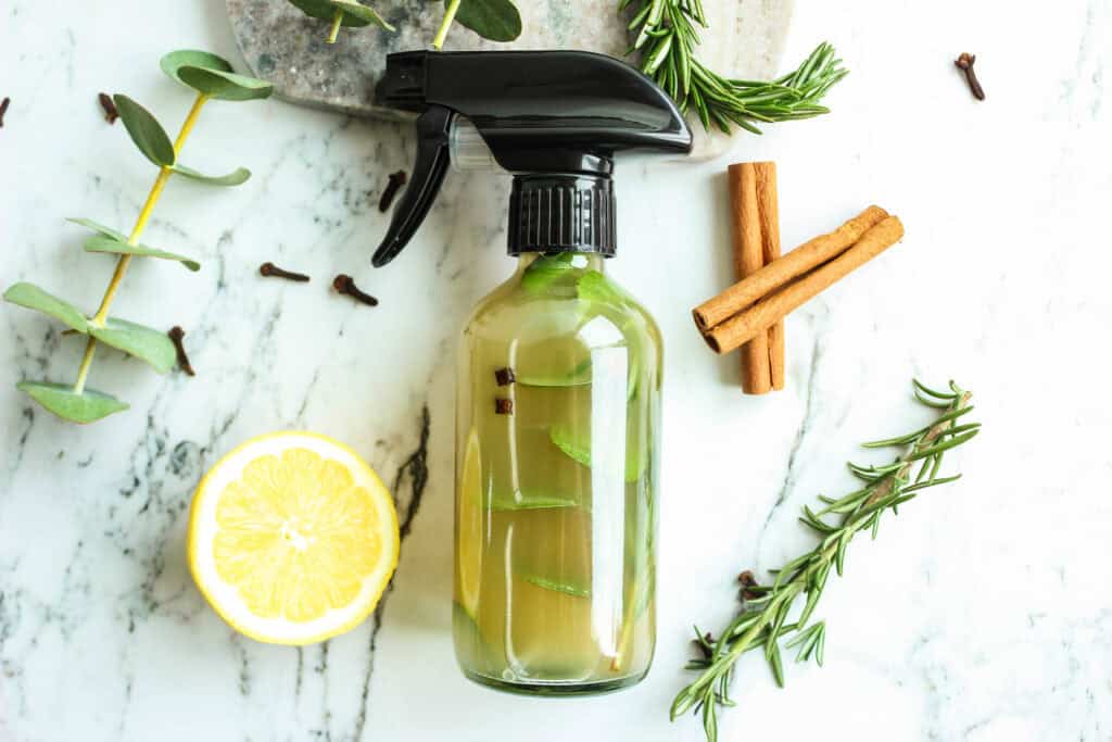 cleaning spray in a clear glass bottle, infused with a cinnamon stick and rosemary sprigs, placed on a marble countertop with lemon slices, whole cloves, and fresh greenery nearby.