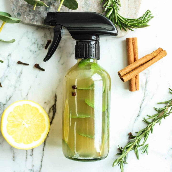 cleaning spray in a clear glass bottle, infused with a cinnamon stick and rosemary sprigs, placed on a marble countertop with lemon slices, whole cloves, and fresh greenery nearby.