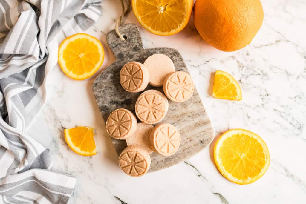 Homemade orange shower steamers on marble with fresh oranges and slices.