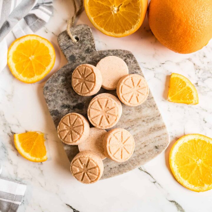 Hand holding a DIY orange shower steamer tablet with sliced citrus in background.