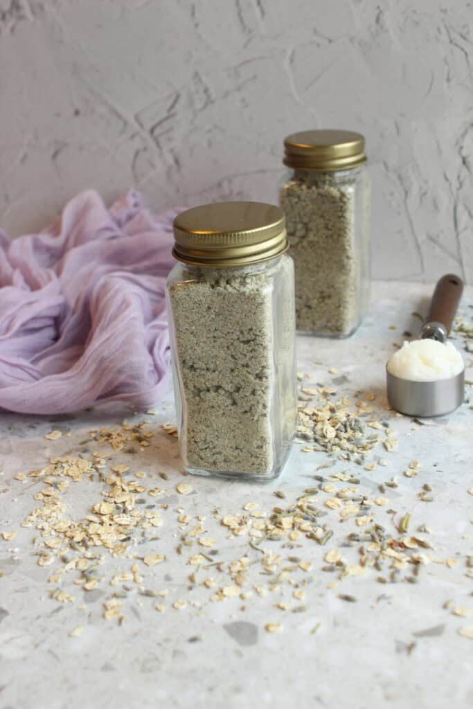 Lavender oatmeal face scrub in glass jars with a scoop on a textured surface.