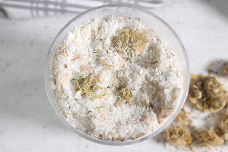Close-up of a glass container holding coarse bath salts mixed with dried botanical pieces.