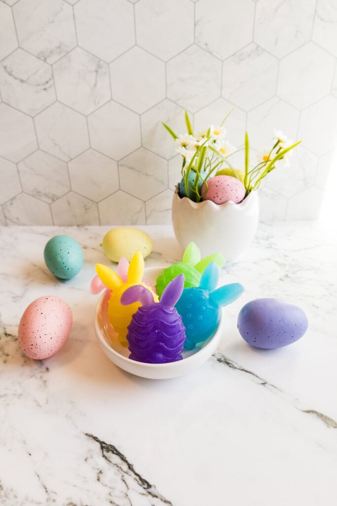 Colorful Easter bunny-shaped soaps with pastel eggs and spring flowers on marble surface.