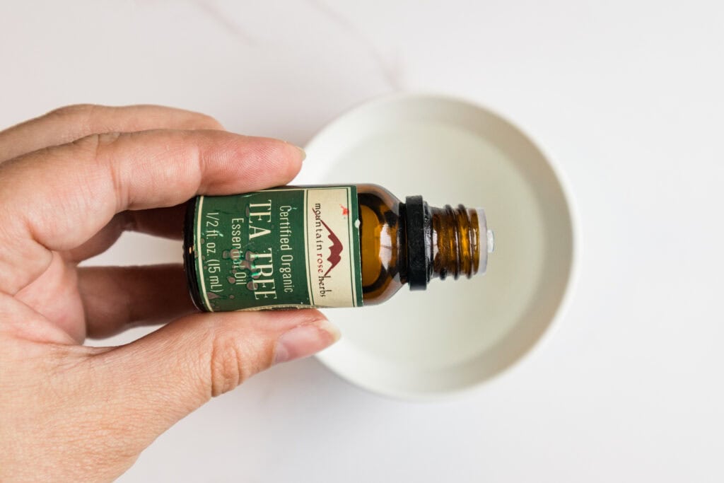Tea Tree Oil bottle held over a small white dish, ready for skincare application.