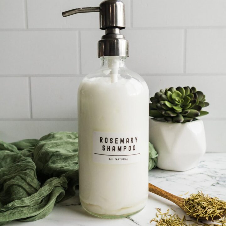 Natural rosemary shampoo in a white dispenser bottle surrounded by green leaves and dried rosemary on a light marble background.