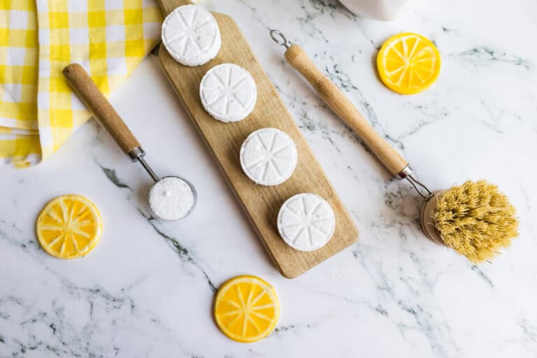 Homemade dishwasher tabs arranged on a wooden board with lemon slices and cleaning tools.