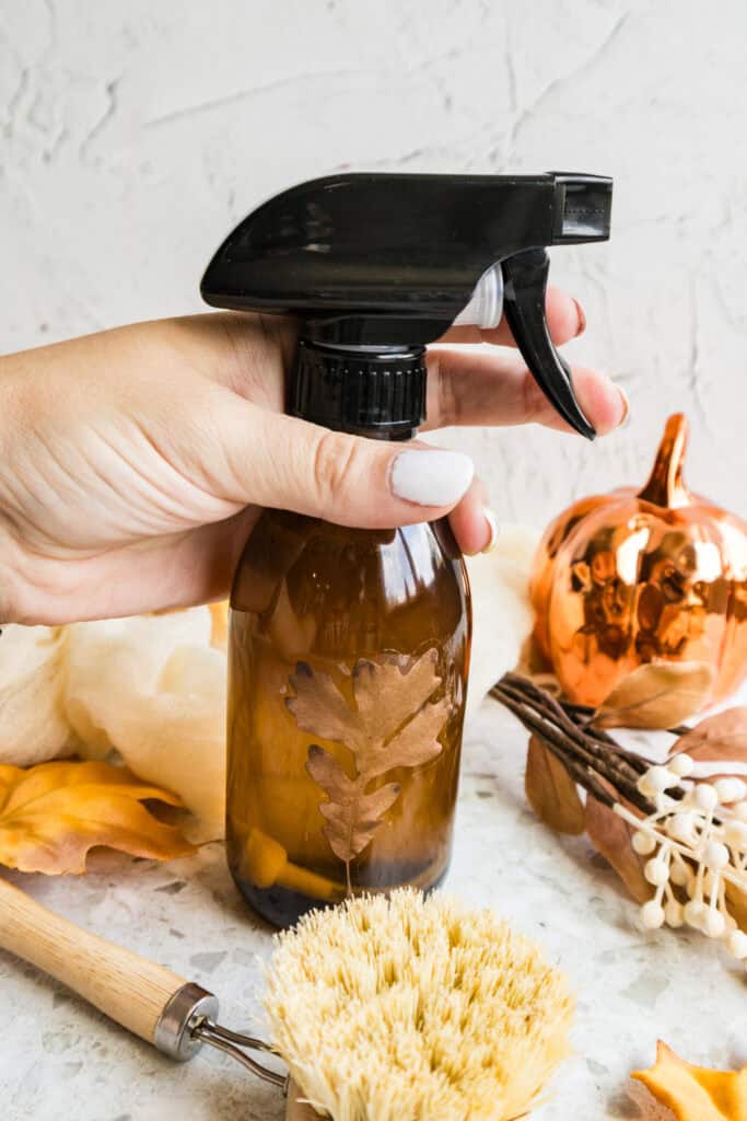 Hand holding an amber trigger spray bottle with autumn decorations.