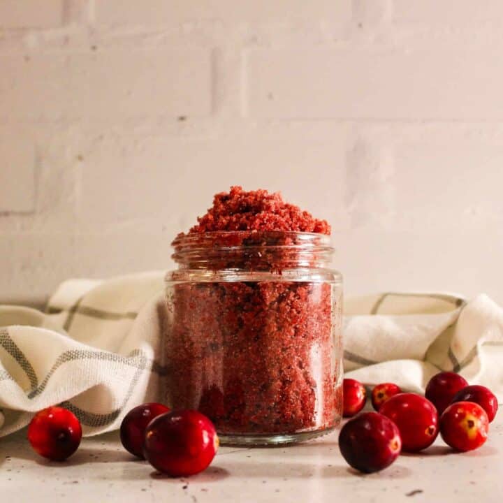 Glass jar filled with red cranberry sugar scrub placed on a cloth with fresh cranberries around it.