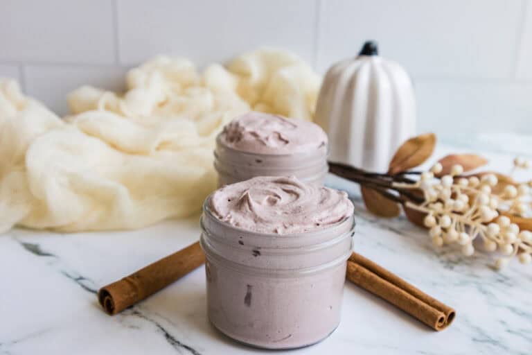 Two open jars of Whipped Tallow Balm with cinnamon sticks and autumn leaves on a marble surface.