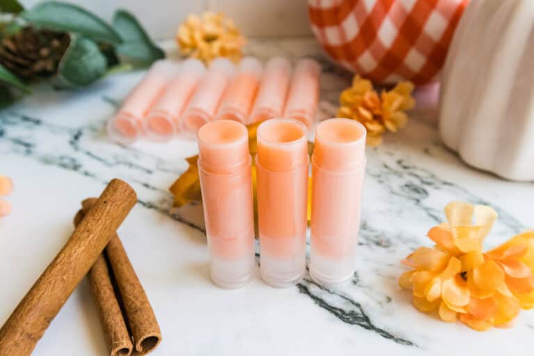 Three orange lip balm tubes displayed with pumpkins, cinnamon sticks, and flowers.