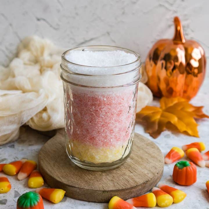 Glass jar filled with layered yellow and pink candy corn bath salts on a wooden coaster with candy corn scattered around.