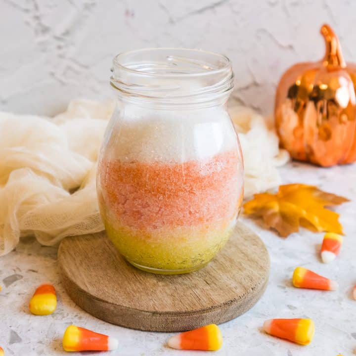 Glass jar of layered yellow, orange, and white sugar scrub on a wooden coaster, surrounded by candy corn and a small orange pumpkin.